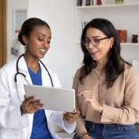 A female doctor consulting with a female patient in a medical examination room, reviewing information on a tablet.